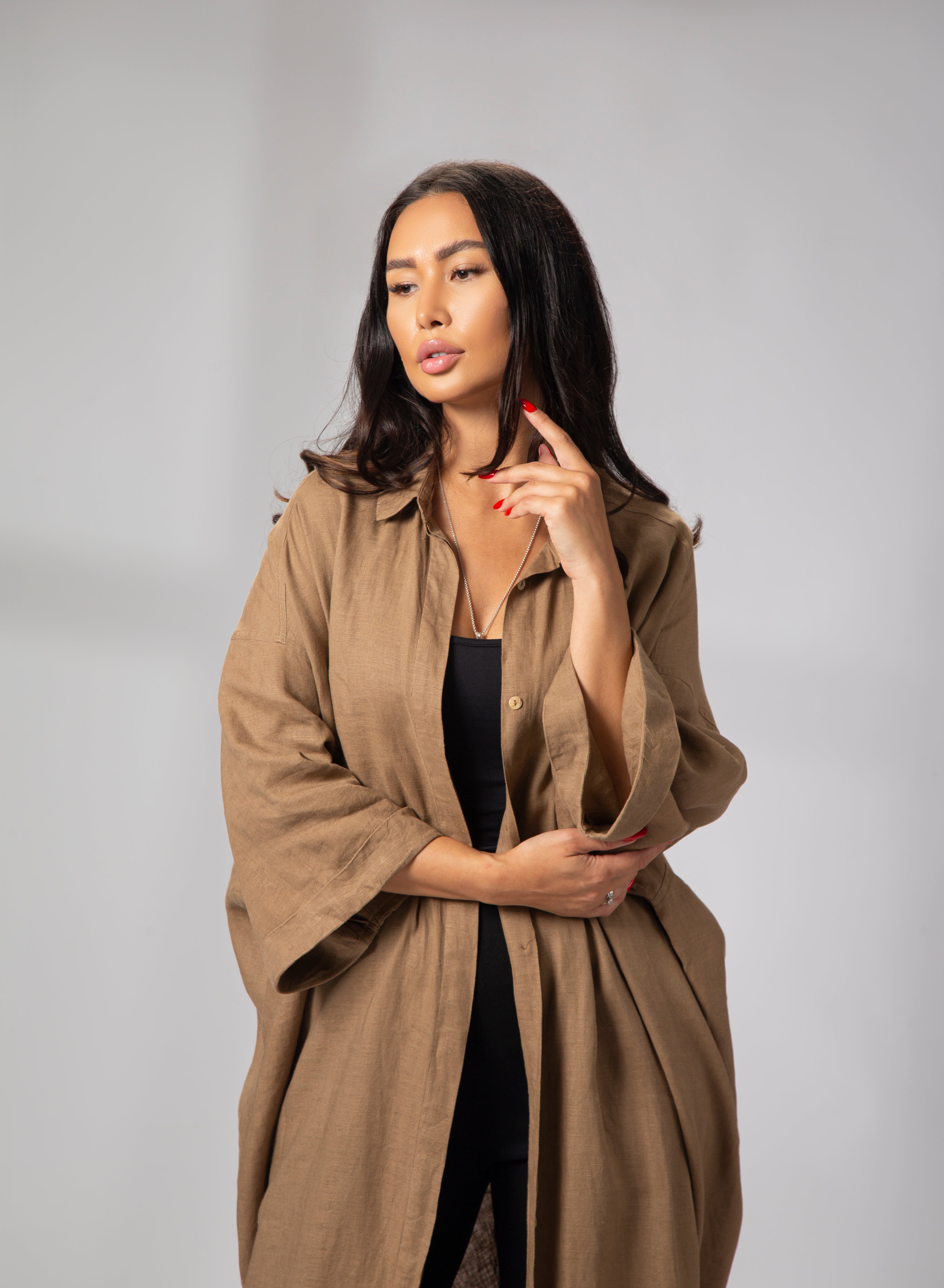 A woman seated on a chair, wearing a long sleeve linen kaftan in camel brown color, with white trousers and light-colored heels. long sleeve linen kaftan camel brown side profile