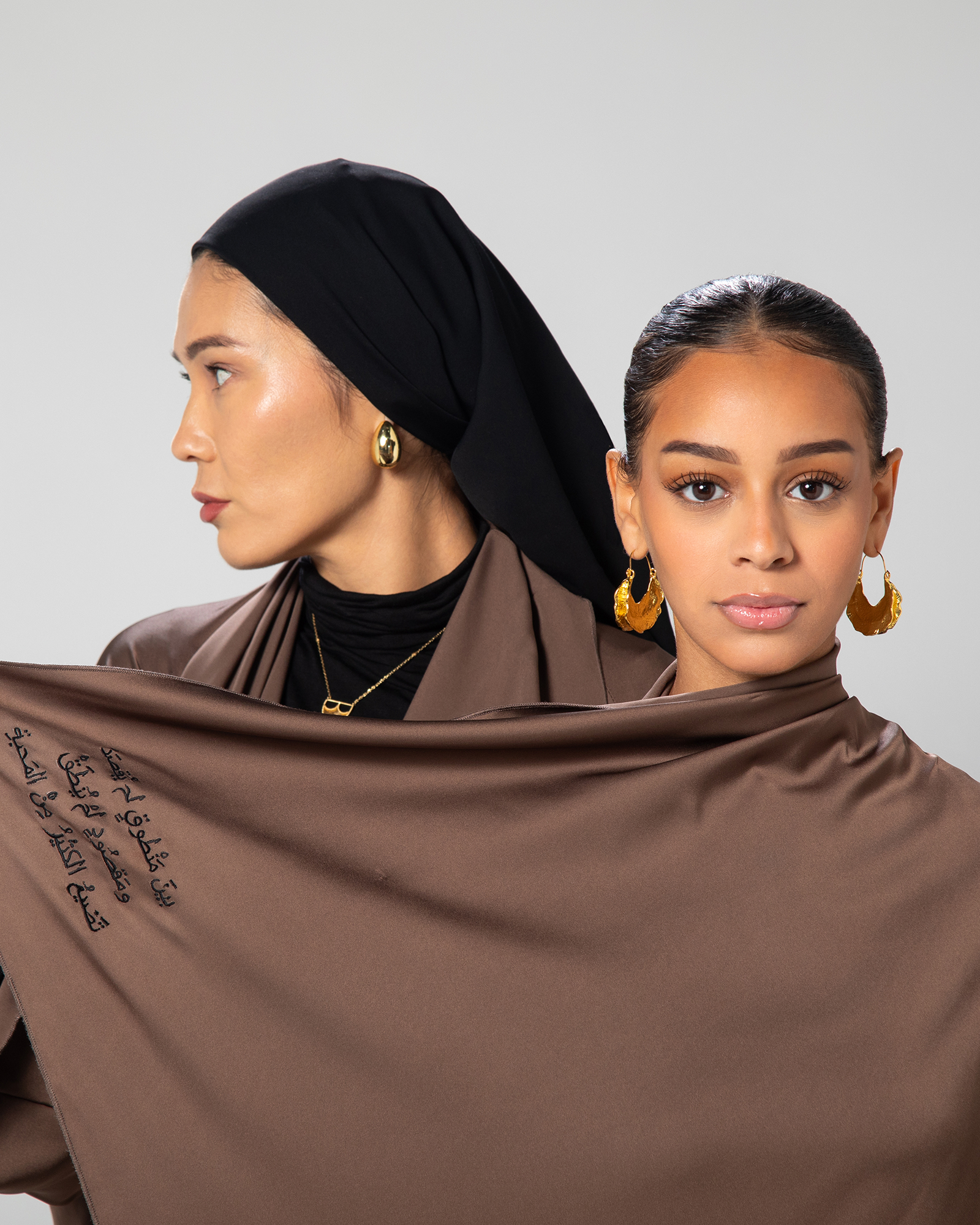 Two women styled in the Flow Abaya by Omni The Label, highlighting the soft brown tone and Arabic calligraphy embroidery across the shoulder KSA UAE Qatar