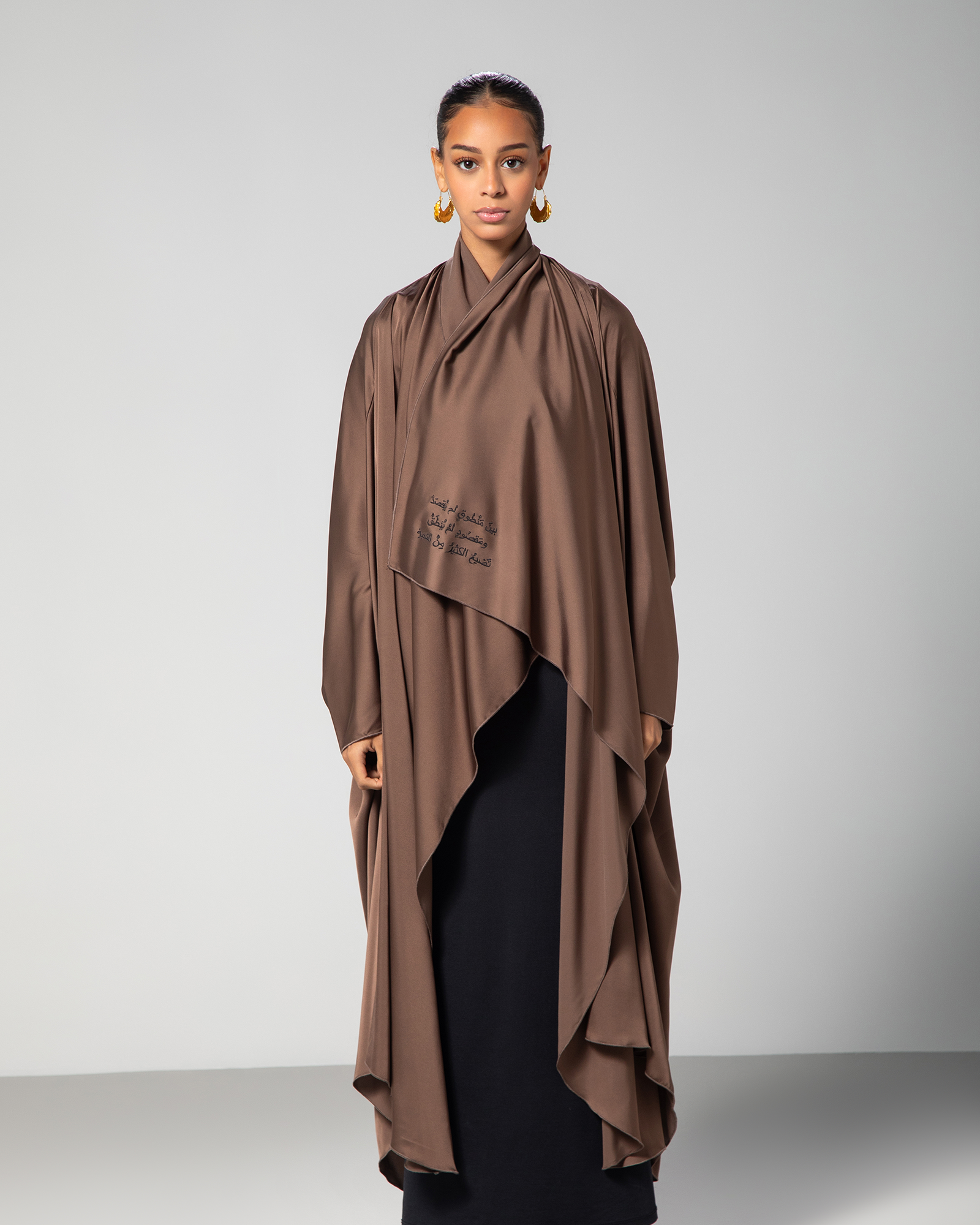 A model wearing a brown silky flow abaya with cascading layers and Arabic calligraphy embroidery on the front.