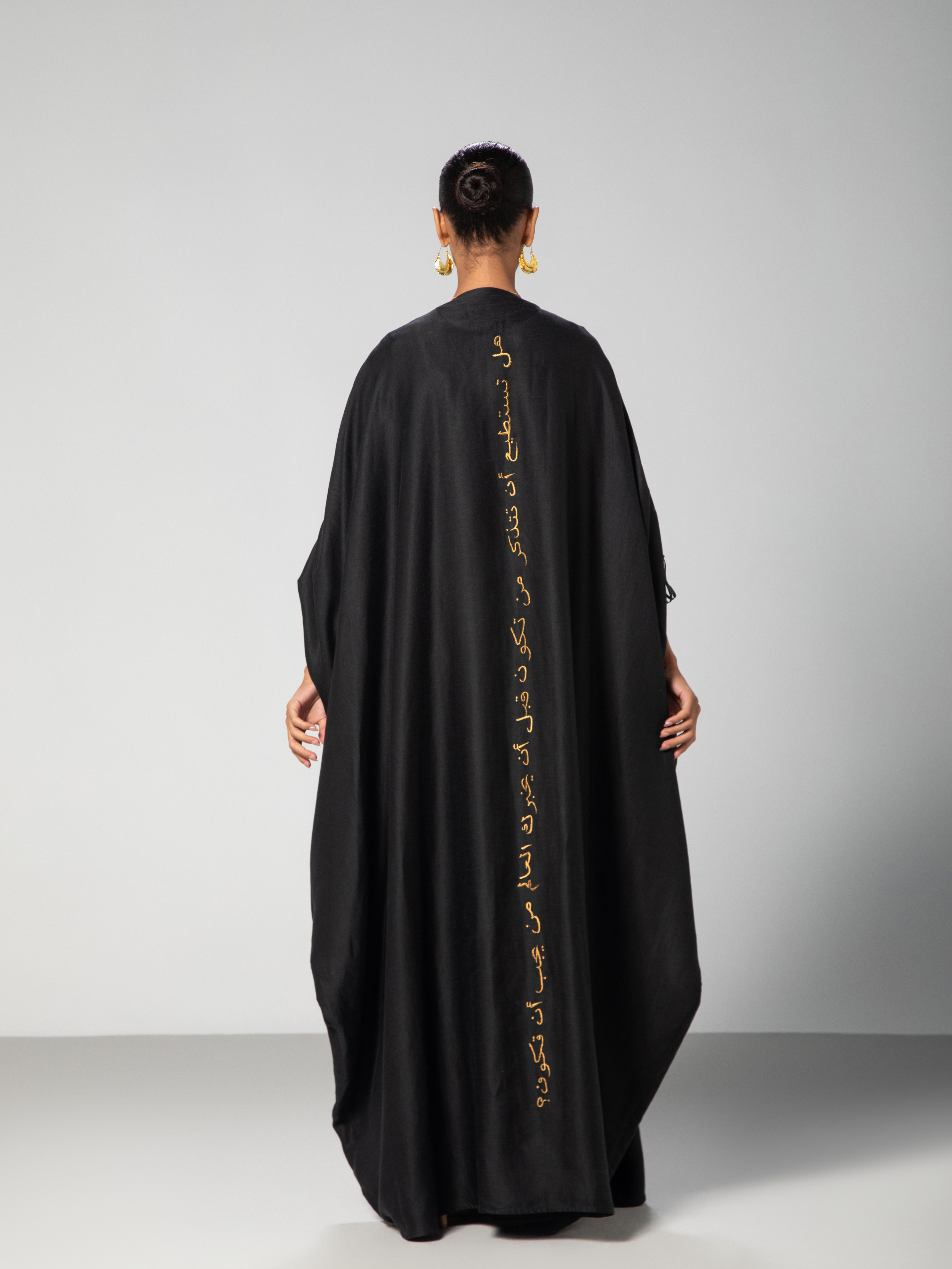 A person facing away from the camera wearing a black linen abaya with gold embroidery on the back and adjustable sleeves UAE KSA Qatar