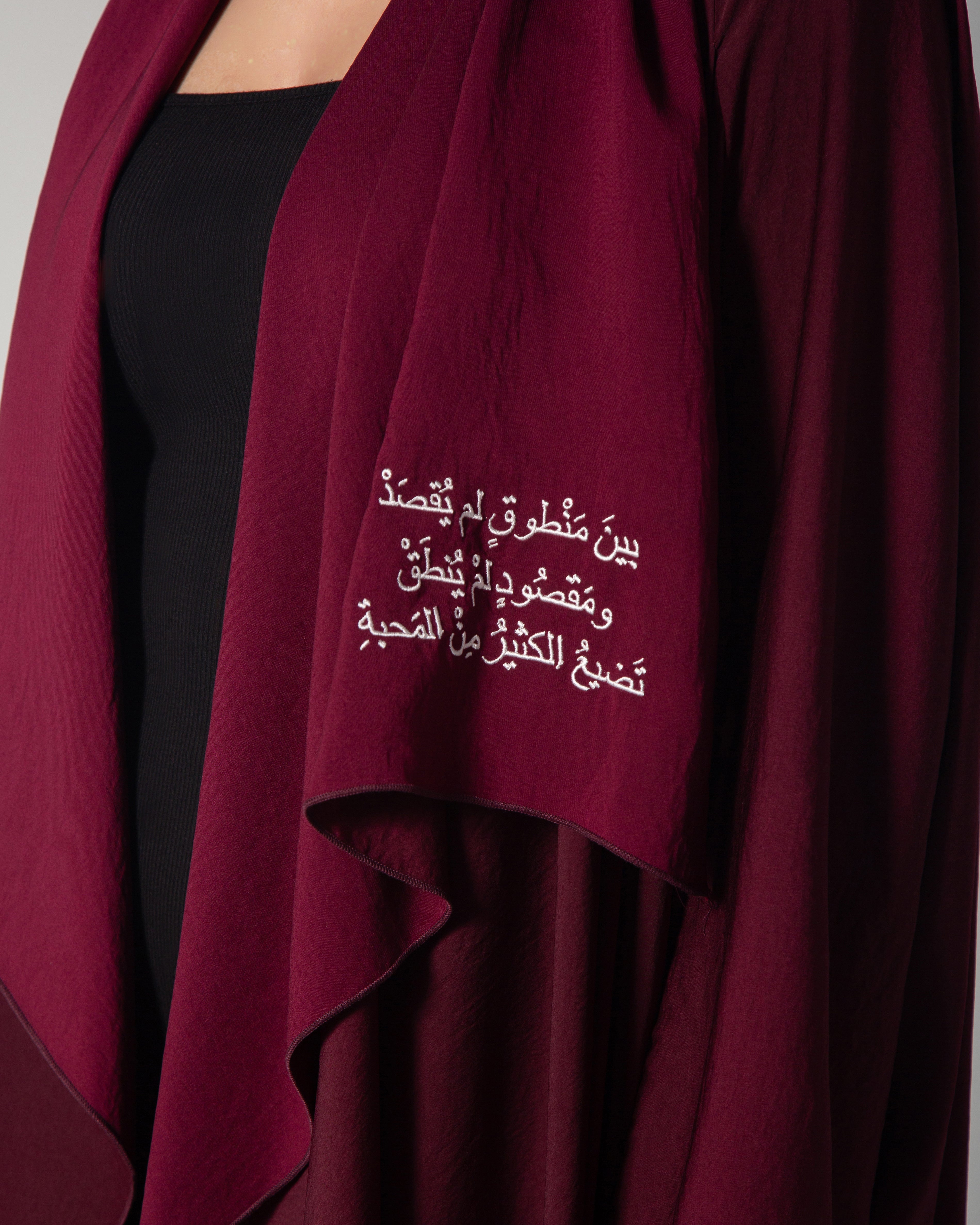 A woman wearing a burgundy abaya with a flowing design and Arabic calligraphy embroidery on the front, middle east saudi kuwait qatar modest fashion