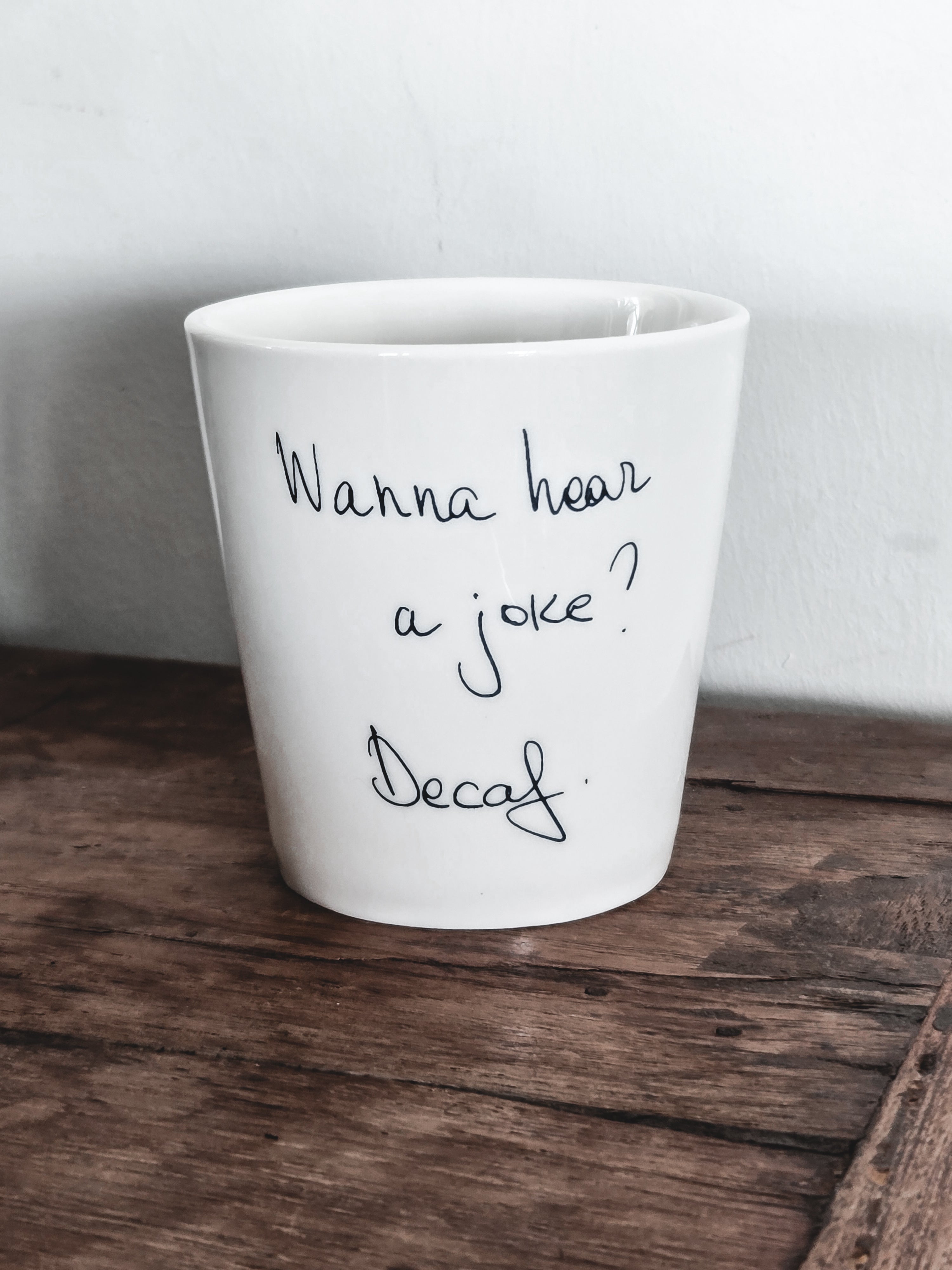 A white ceramic coffee cup with the text 'WANNA HEAR A JOKE? DECAF?' written on it, placed on a wooden surface.