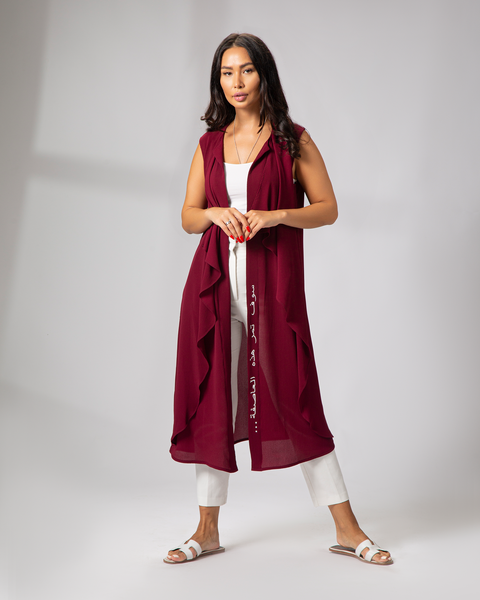A woman standing and posing in a burgundy sleeveless cover-up with embroidery on the front left side, paired with light-colored pants KSA UAE Qatar