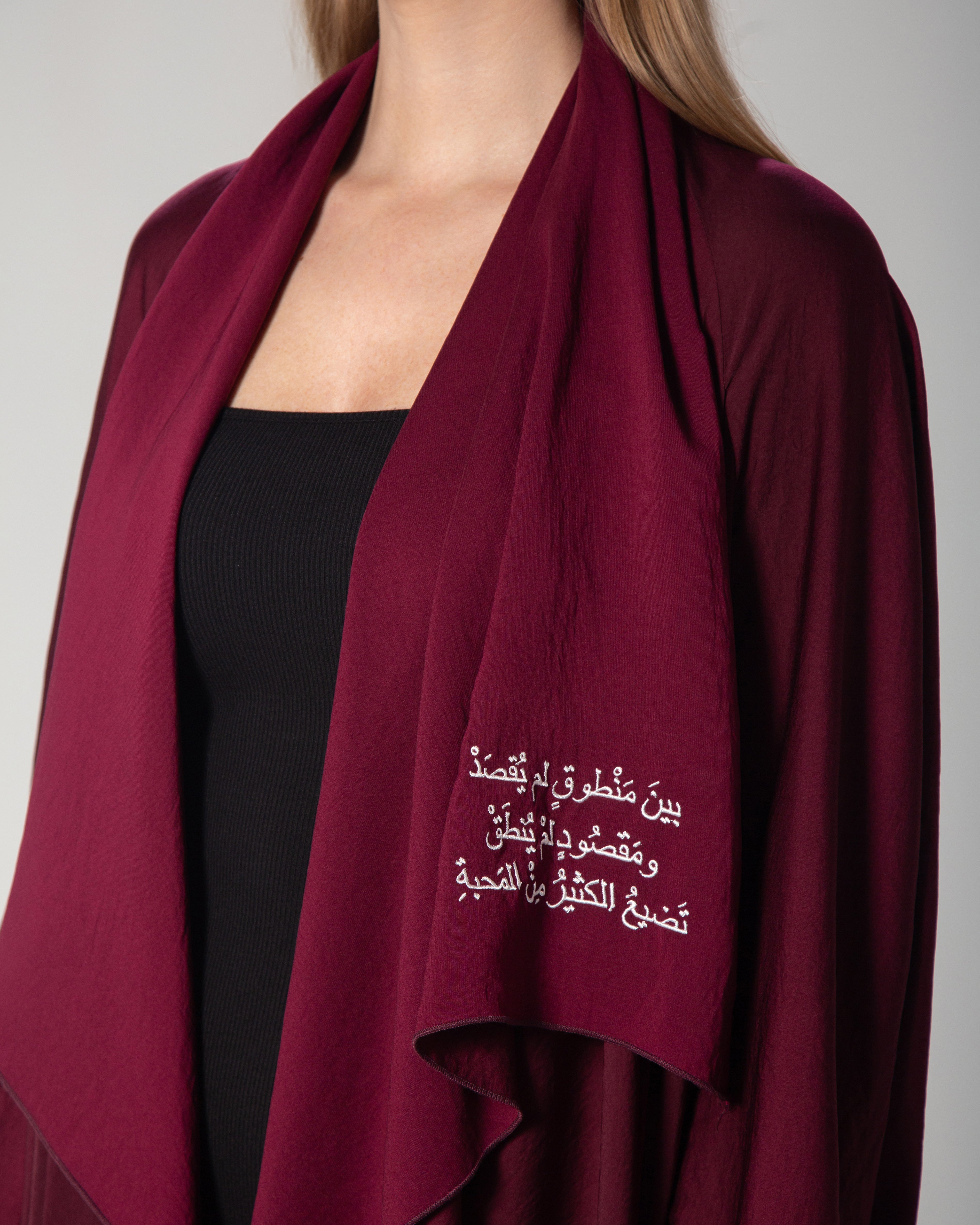 A woman wearing a burgundy abaya with a flowing design and Arabic calligraphy embroidery on the front, middle east saudi kuwait qatar modest fashion omni the label