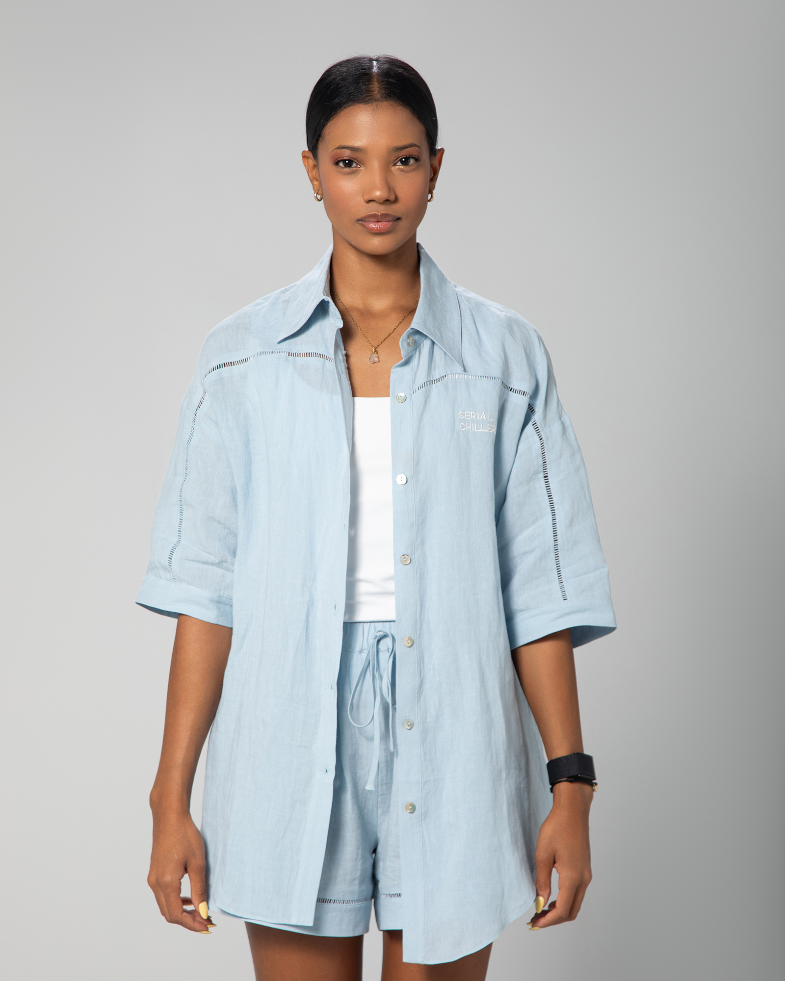Female model wearing a baby blue linen short-sleeve shirt and matching drawstring shorts from Omni The Label, styled with a white tank top for a clean and relaxed summer look UAE KSA Qatar