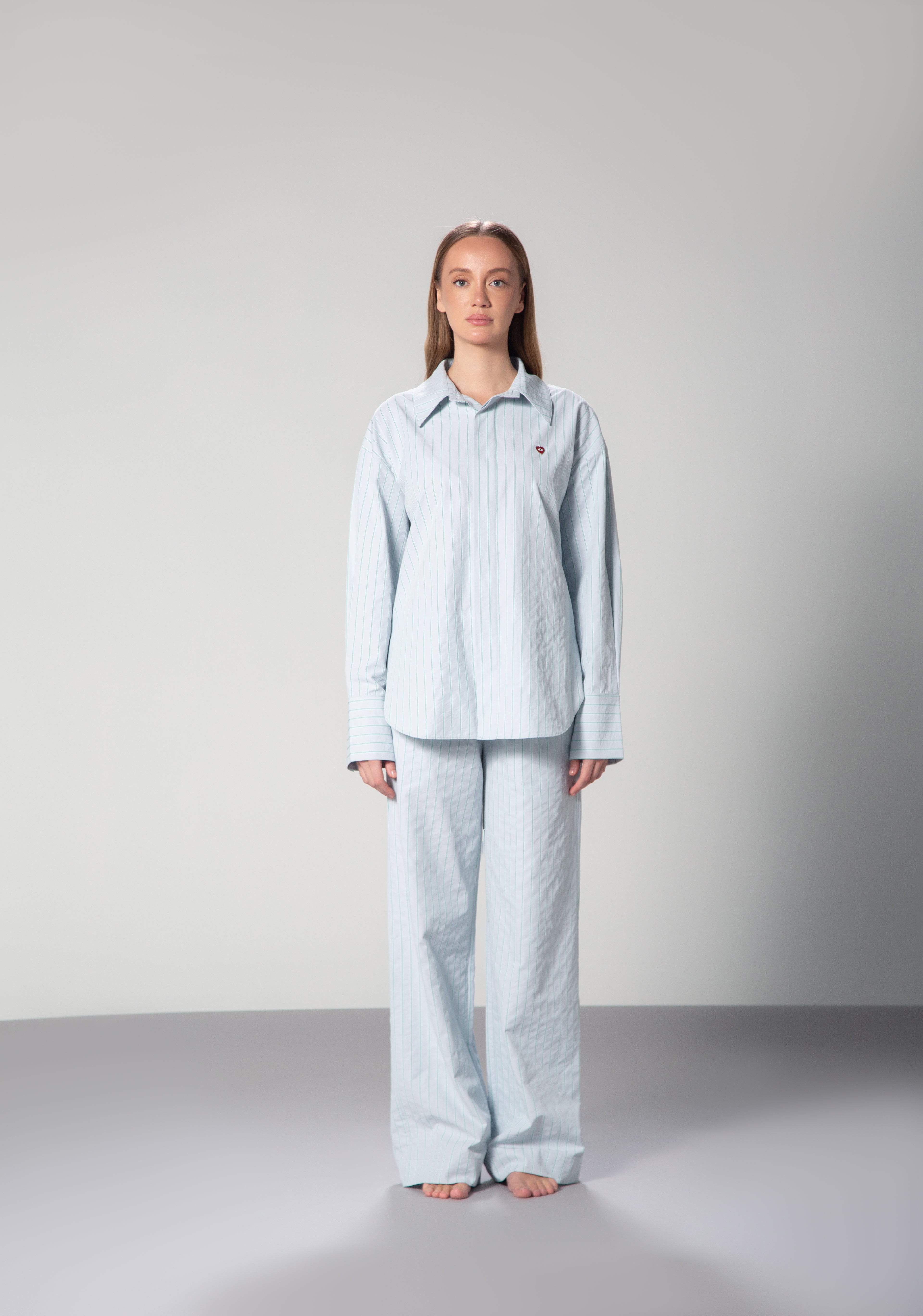 A person standing against a white background, wearing a light blue lounge set with green stripes and a small red heart charm on the front. uae ksa qatar omni the label