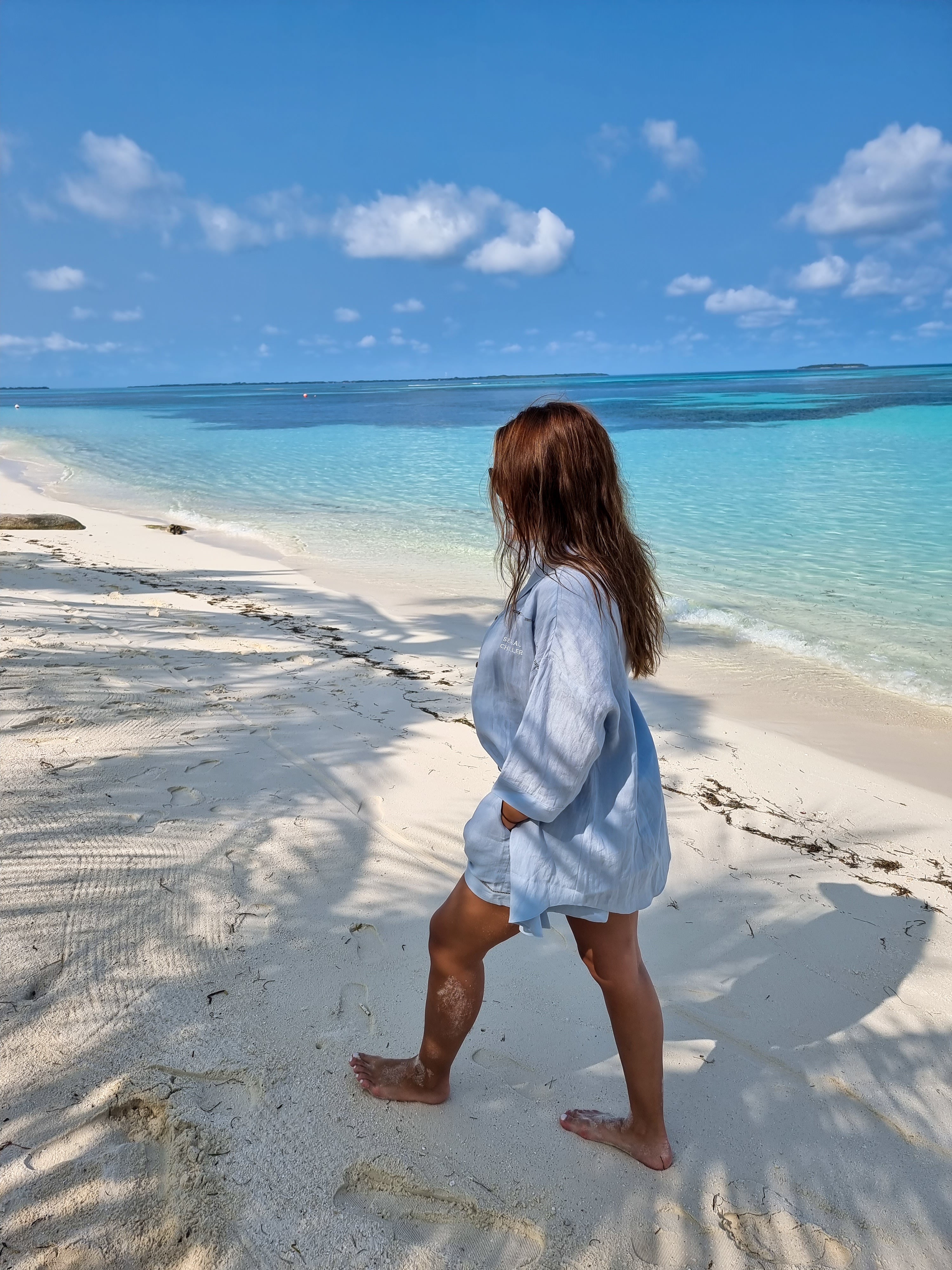 Woman walking barefoot on a tropical beach wearing a light blue linen shirt and shorts set from Omni The Label, showcasing relaxed summer fashion against a turquoise ocean backdrop UAE KSA Qatar