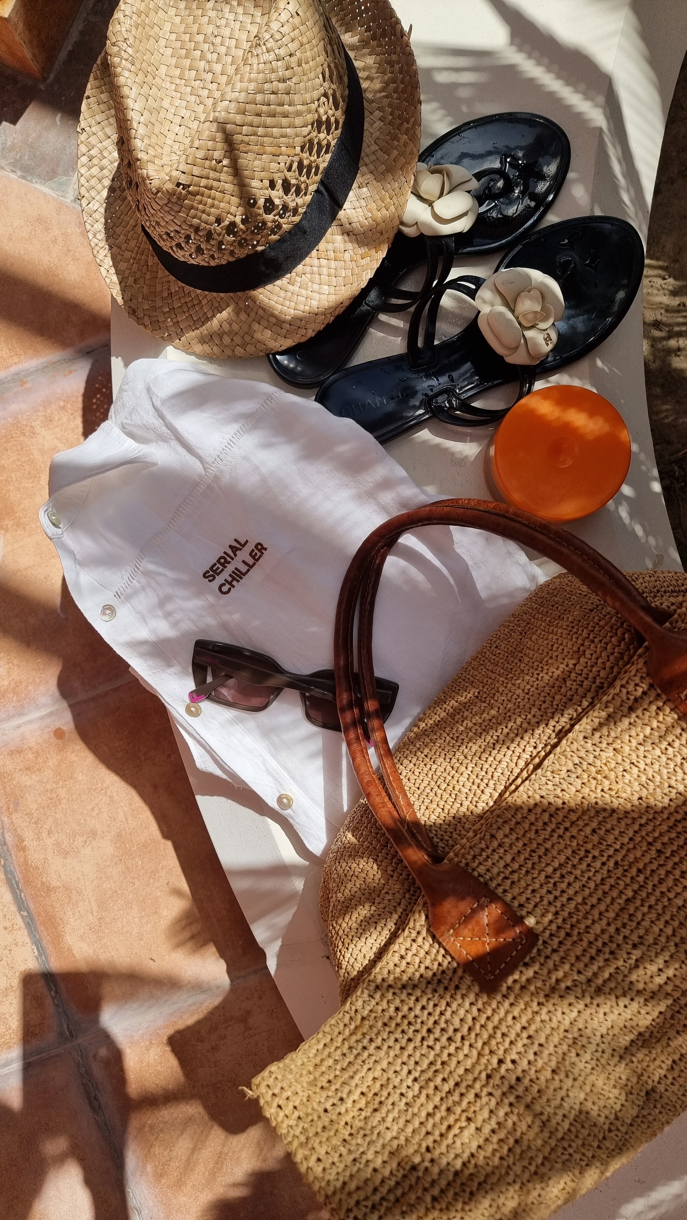 Flat lay of Omni The Label’s white linen shirt with “Serial Chiller” embroidery, styled with a straw hat, sunglasses, black loafers, and a woven beach bag for a summer getaway vibe.UAE, KSA, Qatar