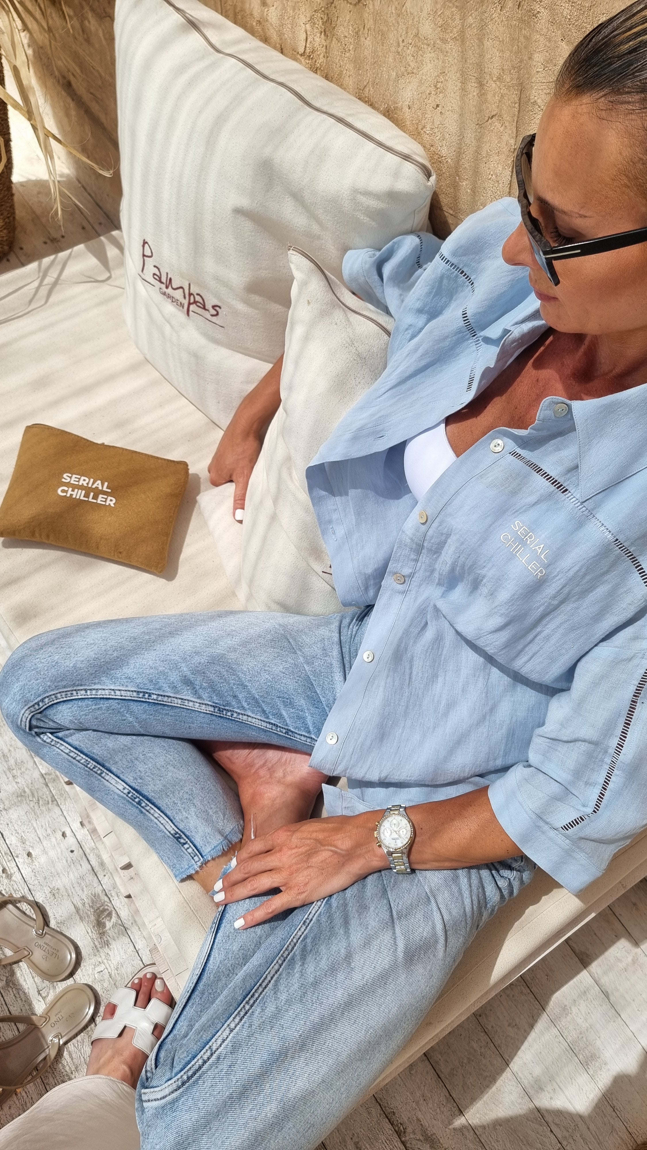 Woman lounging on a neutral-toned couch wearing a light blue linen short-sleeve shirt and matching pants from Omni The Label, accessorized with sunglasses and a gold watch for a casual chic look UAE KSA Qatar