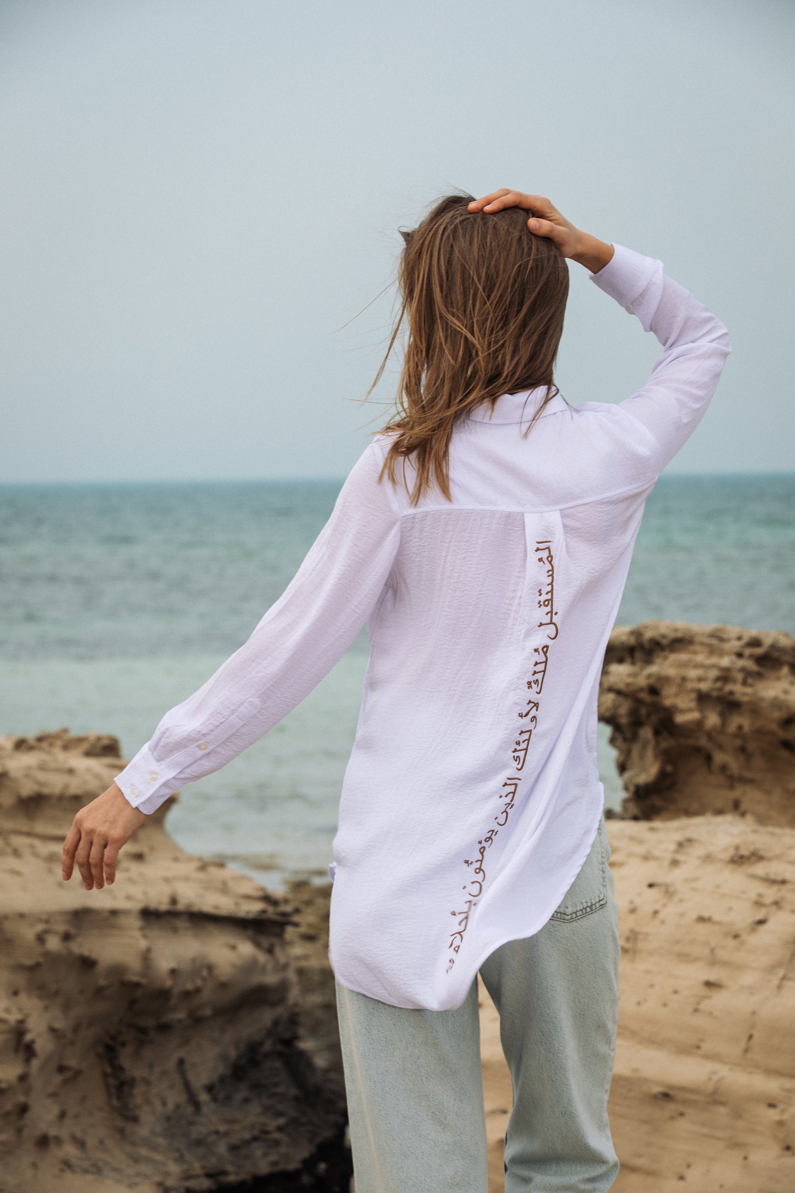 Model at the beach in Omni The Label white shirt catching the wind effortless elegance for modern women UAE Qatar KSA