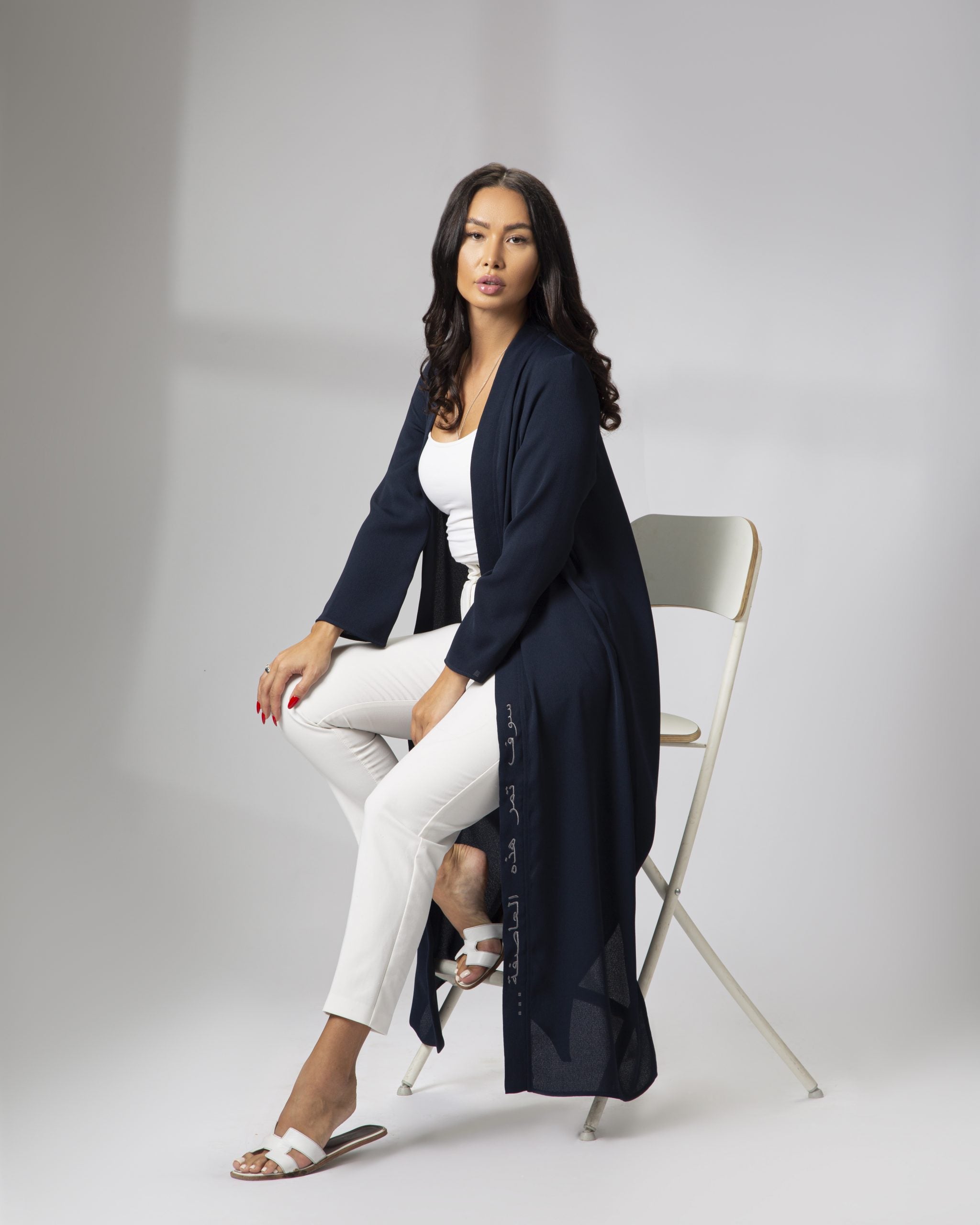Model seated in a white chair wearing Omni The Label’s navy blue kimono with Arabic embroidery, styled over a white outfit and sandals KSA UAE Qatar