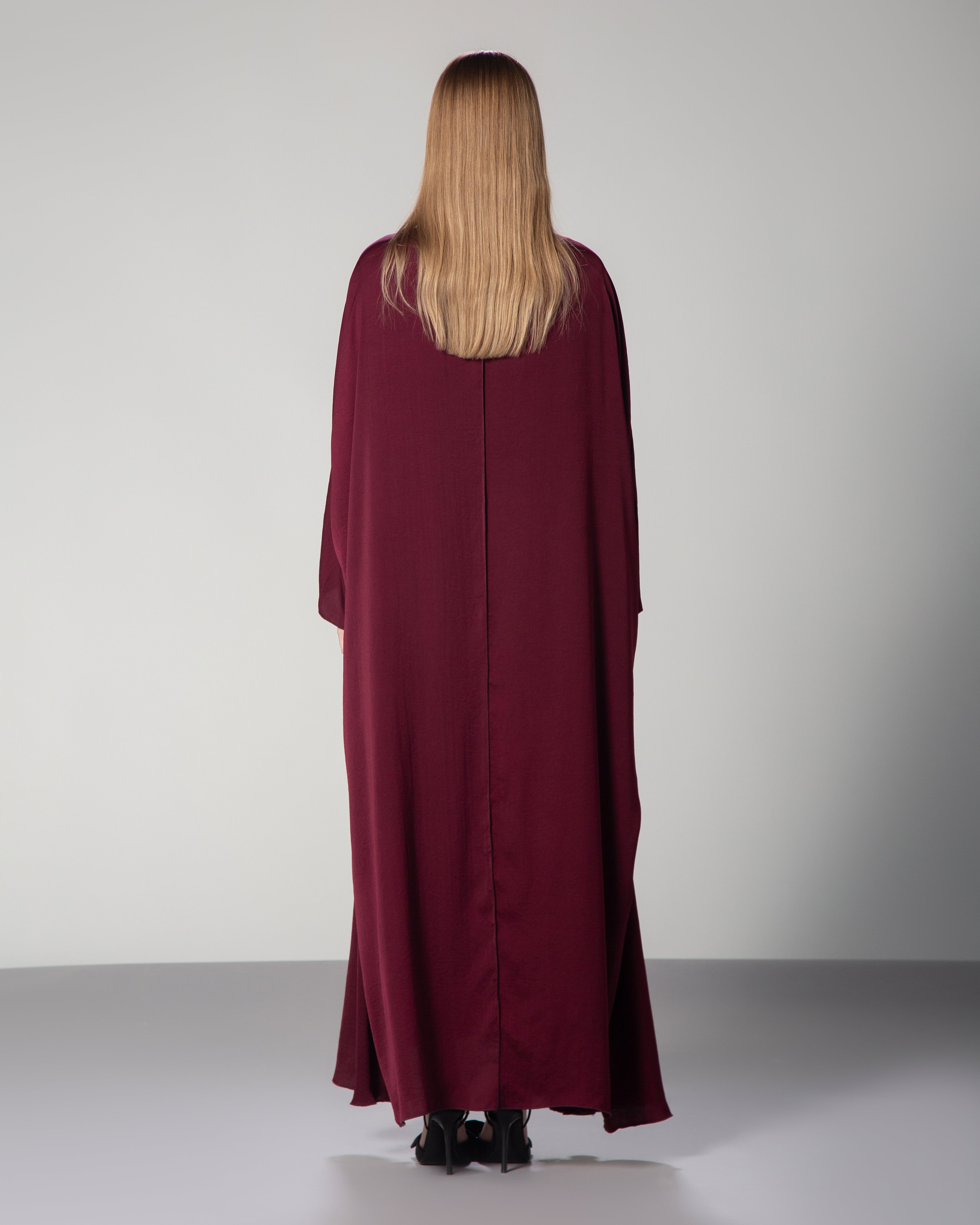 A woman wearing a burgundy abaya with a flowing design and Arabic calligraphy embroidery on the front. saudi qatar kuwait fashion omni the label 