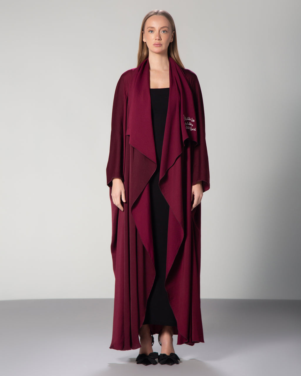 A woman wearing a burgundy abaya with a flowing design and Arabic calligraphy embroidery on the front.Between what is said and not meant, and what was meant but not said, most love is lost." saudi qatar dubai kuwait fashion 