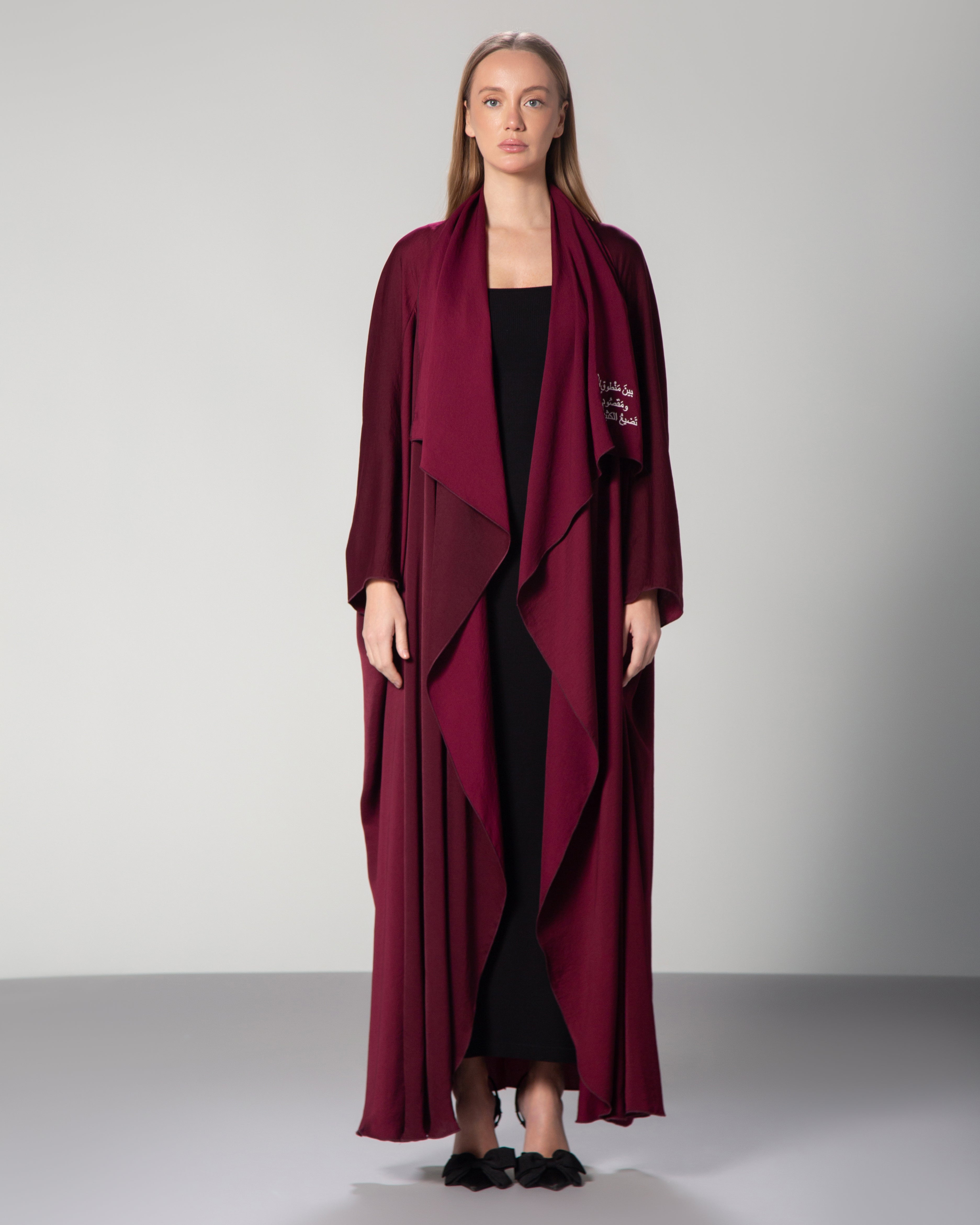 A woman wearing a burgundy abaya with a flowing design and Arabic calligraphy embroidery on the front.Between what is said and not meant, and what was meant but not said, most love is lost." saudi qatar dubai kuwait fashion 