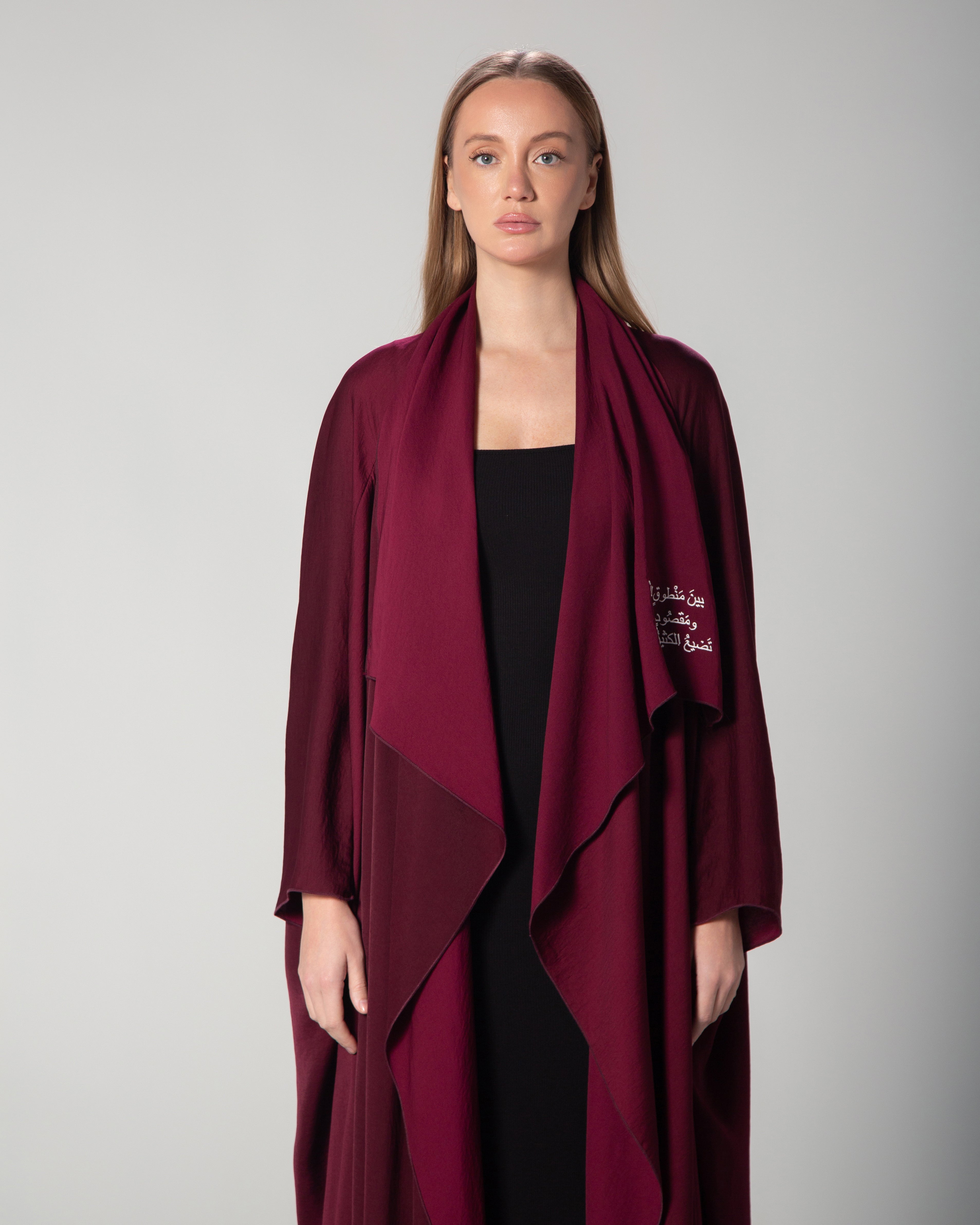 A woman wearing a burgundy abaya with a flowing design and Arabic calligraphy embroidery on the front. saudi qatar kuwait fashion omni the label 