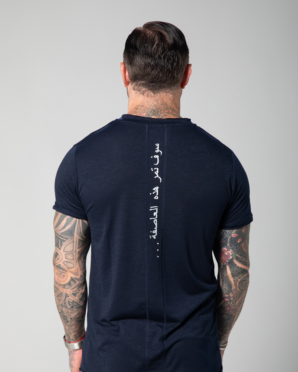The image shows the back view of a male model wearing a navy blue V-neck t-shirt with white text embroidery on the shoulder UAE KSA Qatar