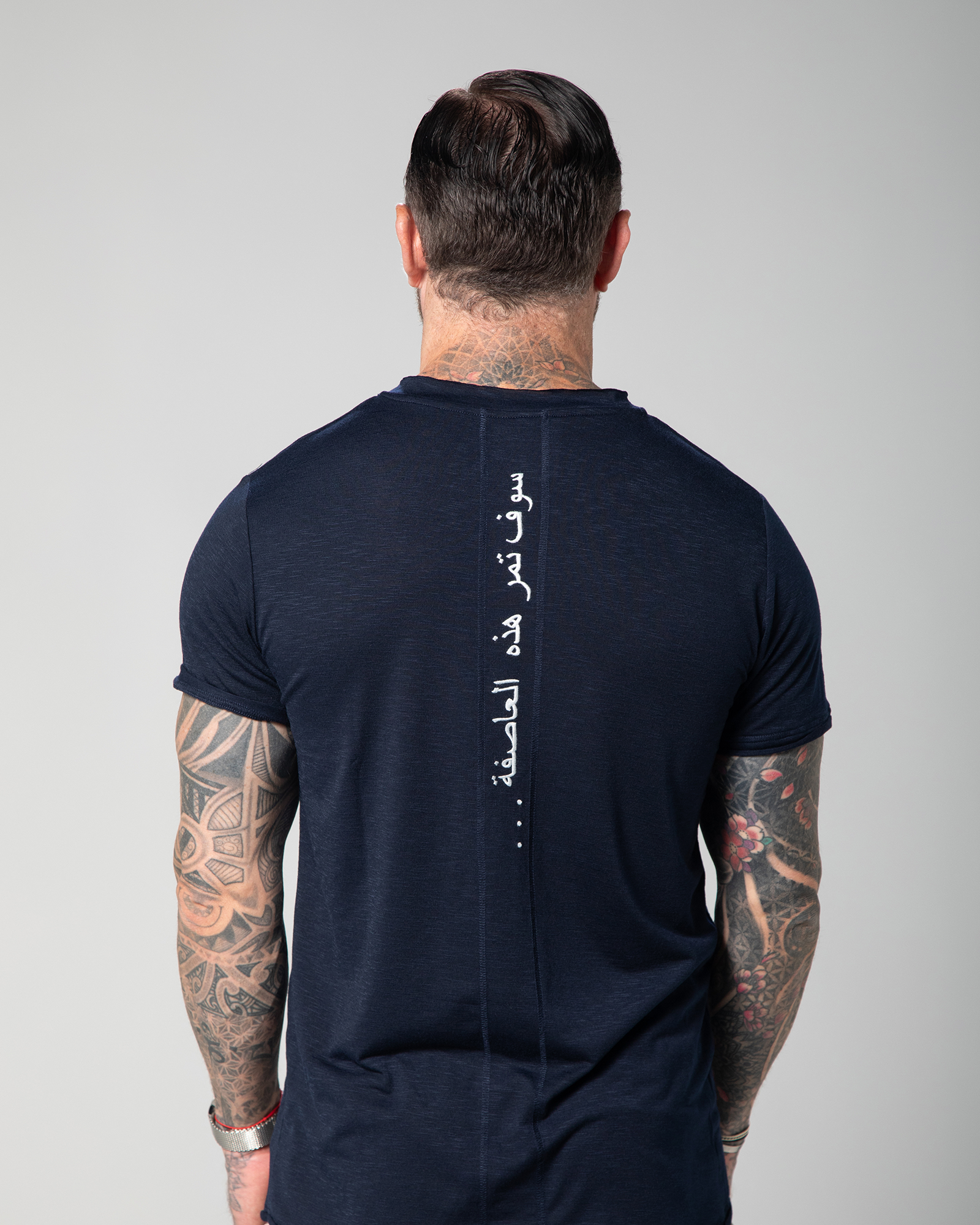 The image shows the back view of a male model wearing a navy blue V-neck t-shirt with white text embroidery on the shoulder UAE KSA Qatar