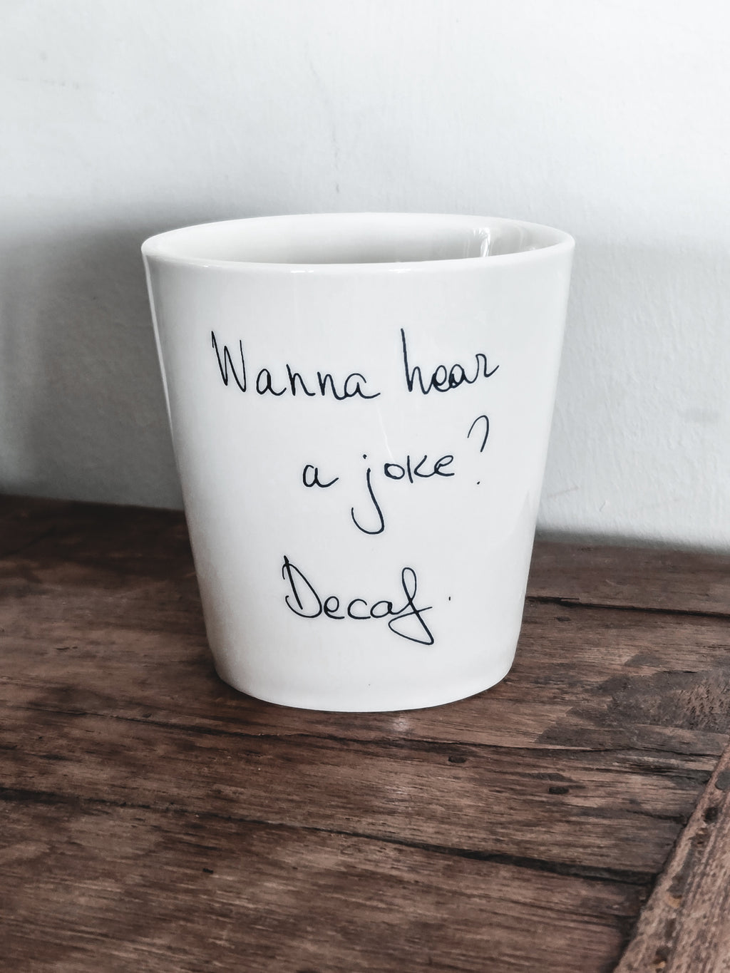 A white ceramic coffee cup with the text 'WANNA HEAR A JOKE? DECAF?' written on it, placed on a wooden surface.