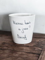 A white ceramic coffee cup with the text 'WANNA HEAR A JOKE? DECAF?' written on it, placed on a wooden surface.