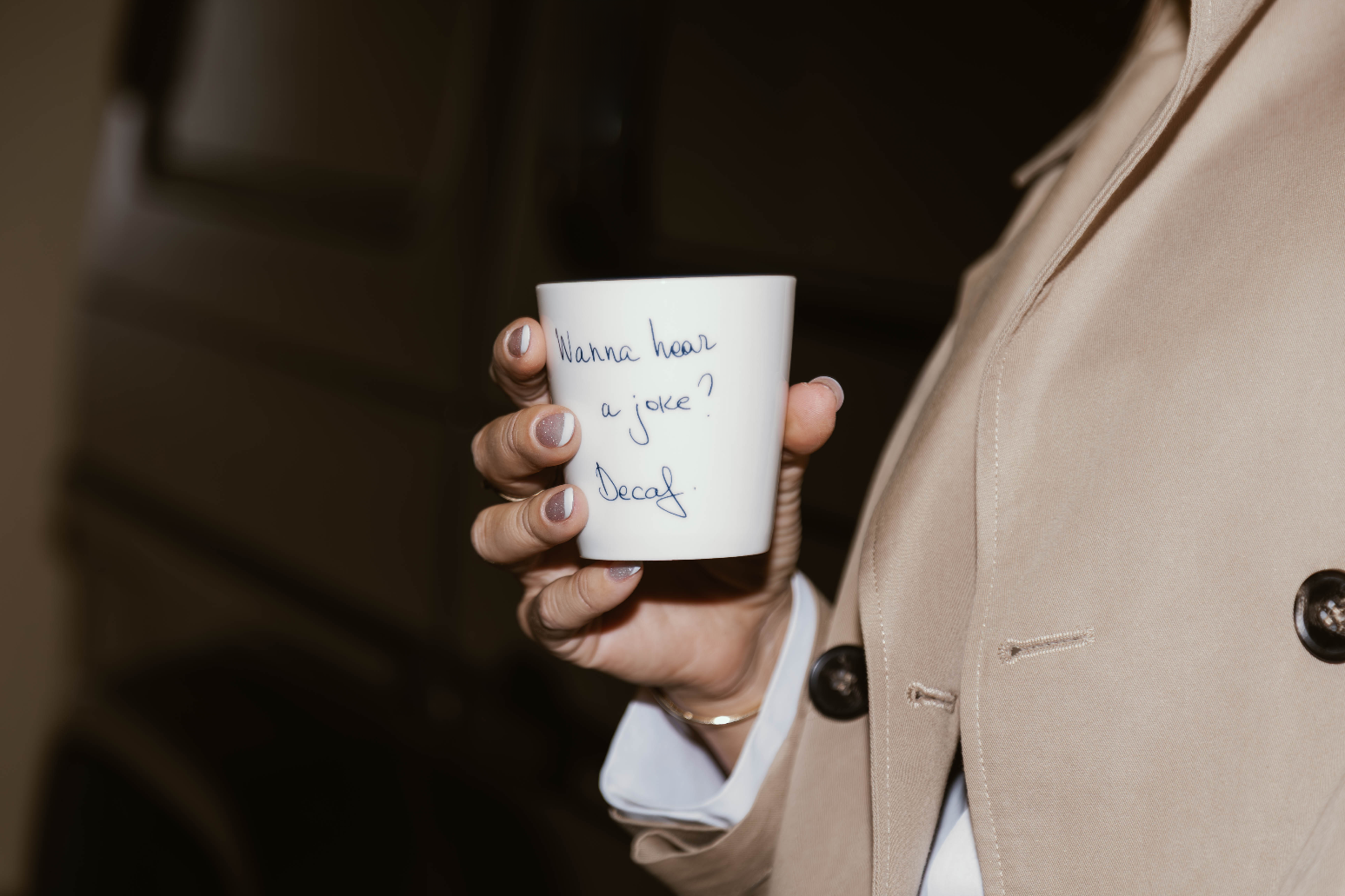 Person holding a white mug with text in a beige coat against a blurred background