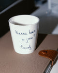 A white china coffee cup with the text 'Wanna hear a joke? Decaf' written on it, displayed on a brown surface, possibly a table or desk.