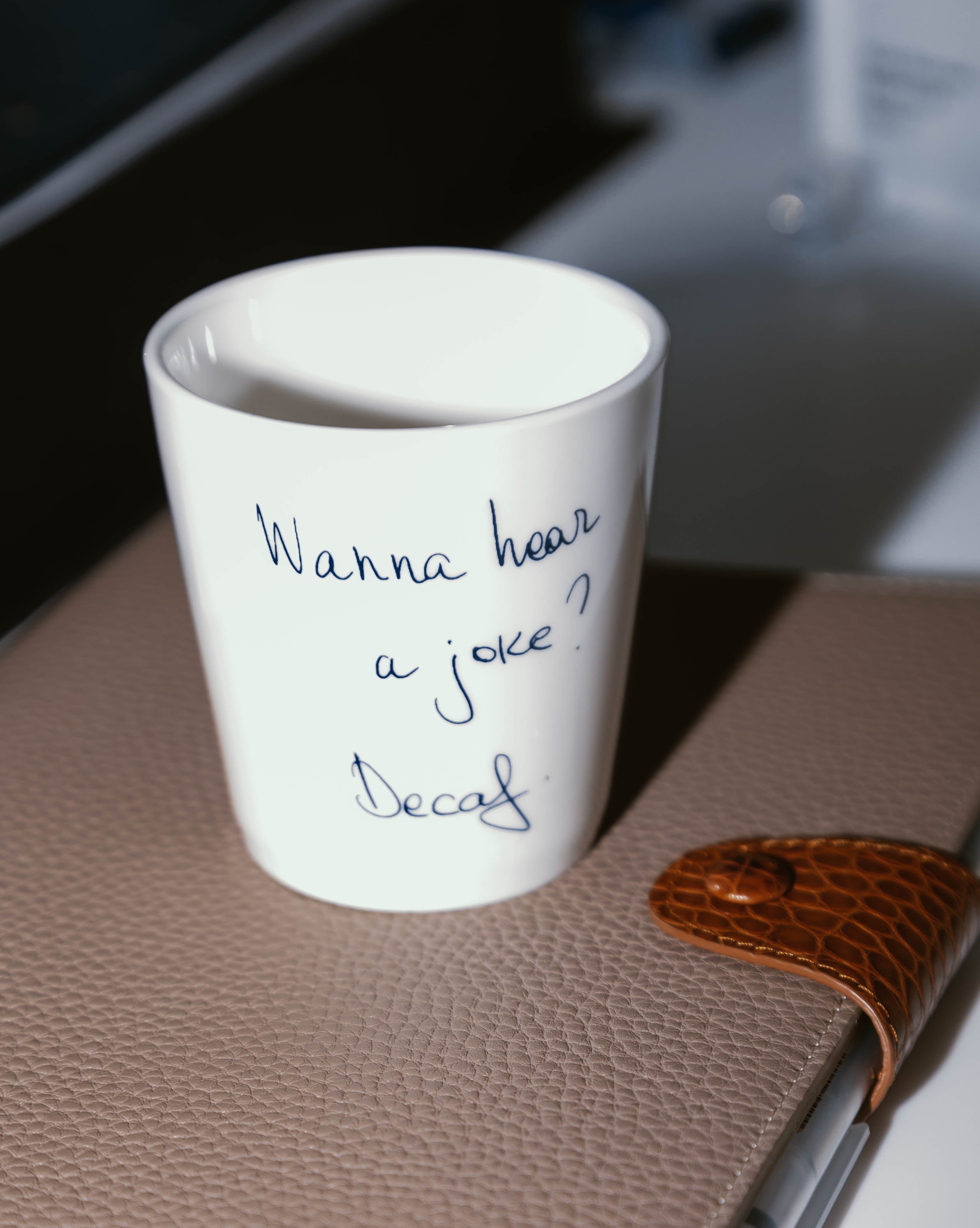 A white china coffee cup with the text 'Wanna hear a joke? Decaf' written on it, displayed on a brown surface, possibly a table or desk.