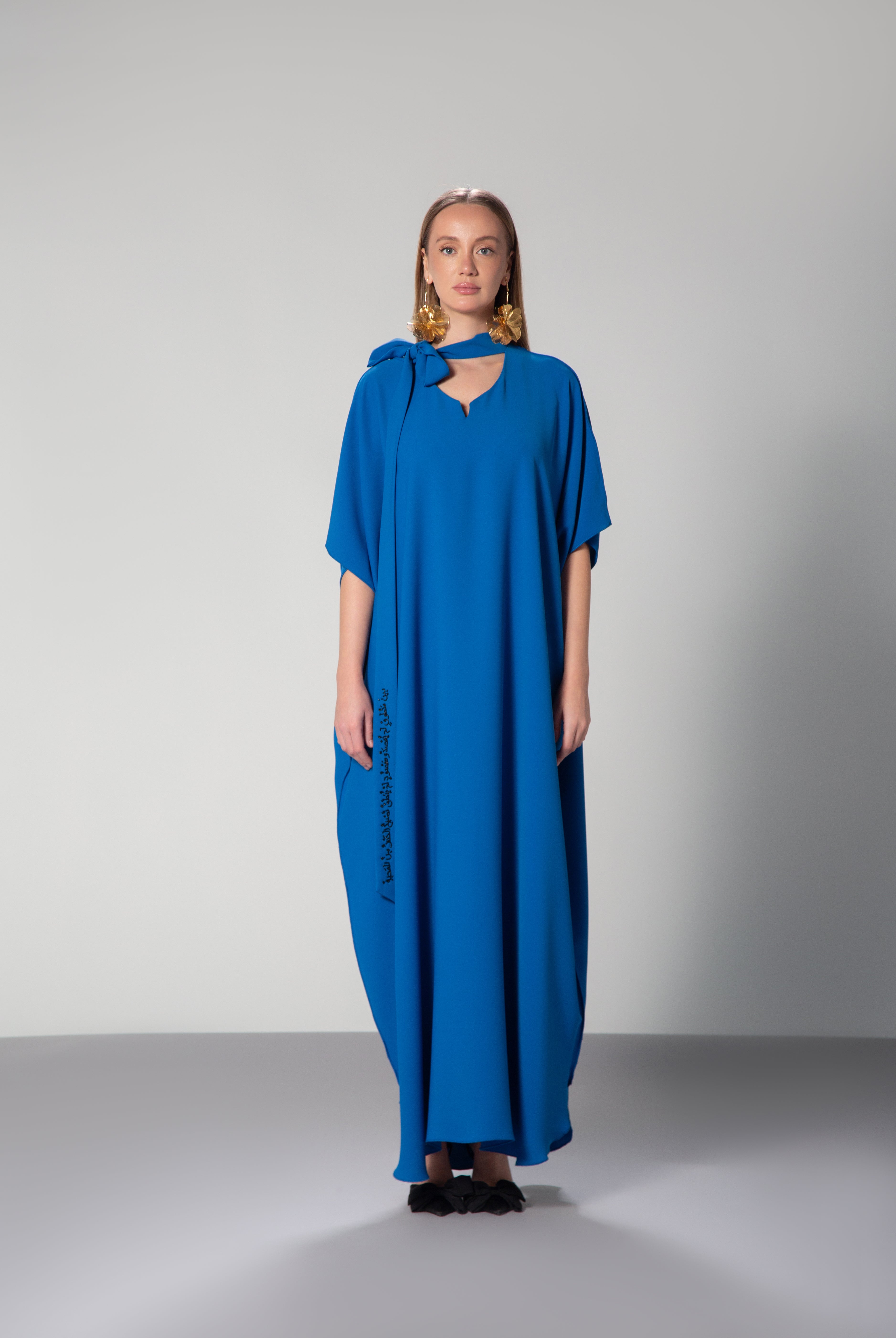 A model wearing a long royal blue dress with half sleeves and a necktie, featuring black embroidery and two slits on each side KSA UAE Qatar