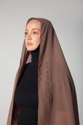 A woman wearing a brown scarf with black embroidery that includes text, paired with a black outfit KSA UAE Qatar