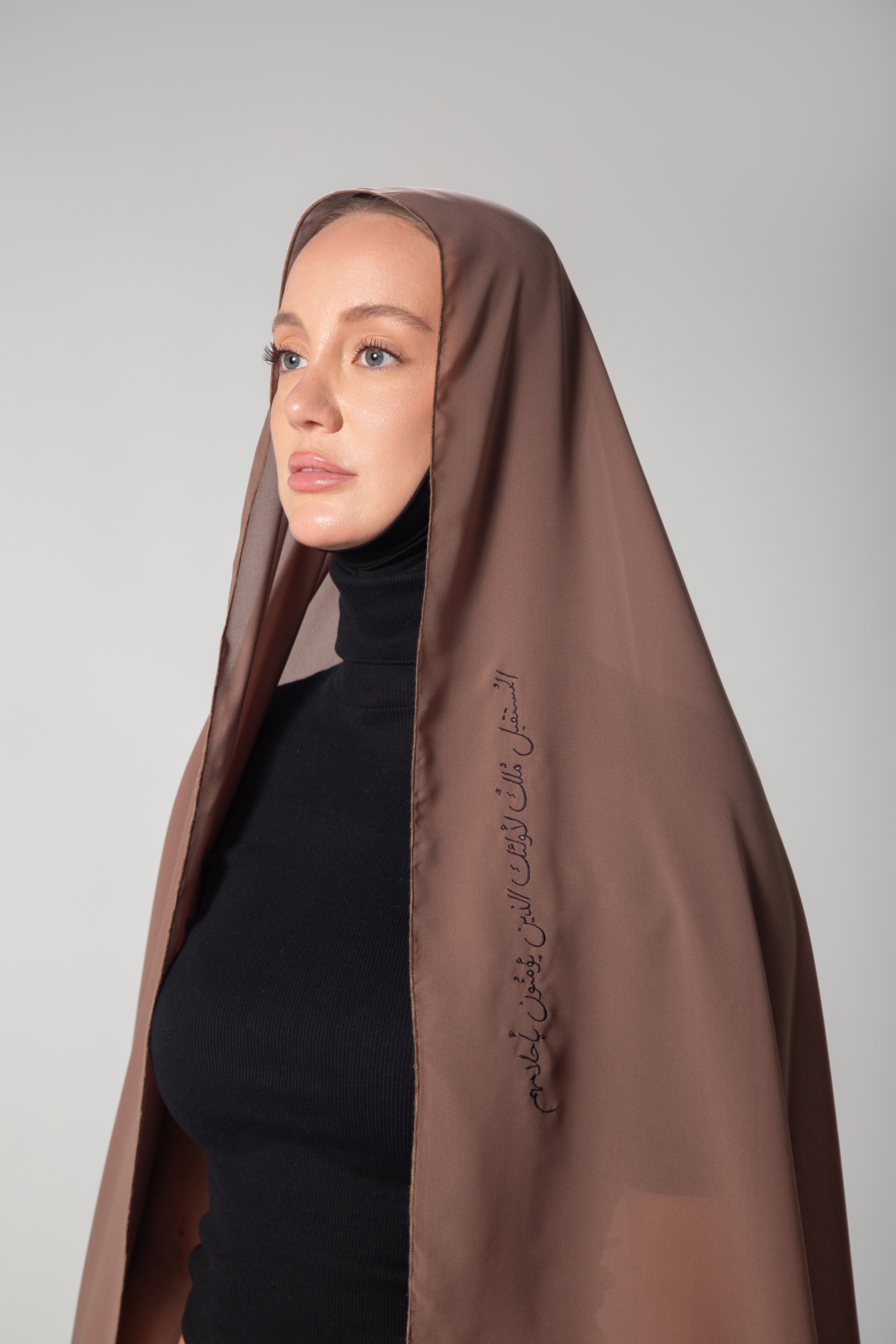A woman wearing a brown scarf with black embroidery that includes text, paired with a black outfit KSA UAE Qatar
