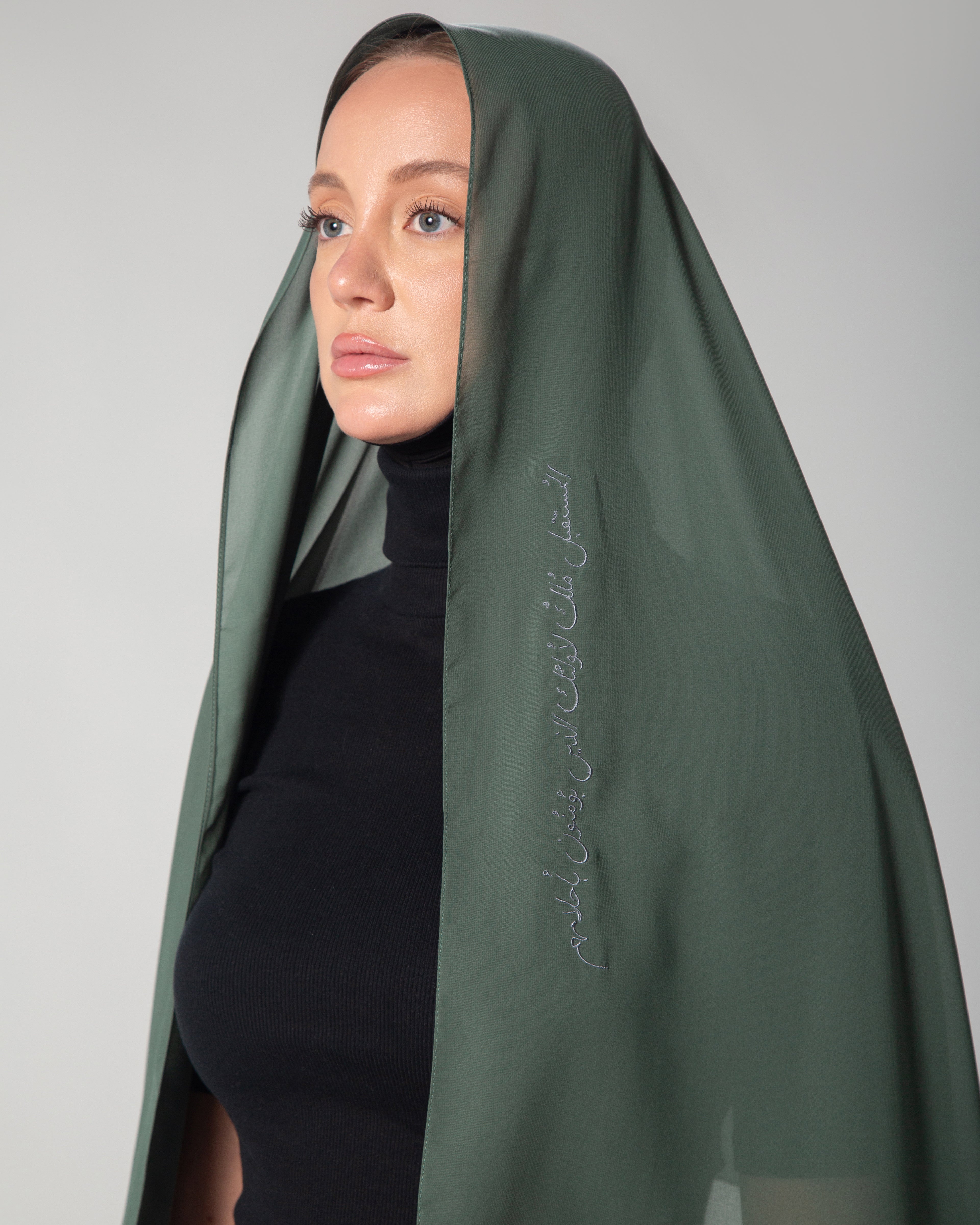 A woman wearing a light green scarf with dark gray embroidery that includes text, paired with a black outfit KSA UAE Qatar