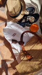 Flat lay of Omni The Label’s white linen shirt with “Serial Chiller” embroidery, styled with a straw hat, sunglasses, black loafers, and a woven beach bag for a summer getaway vibe.UAE, KSA, Qatar