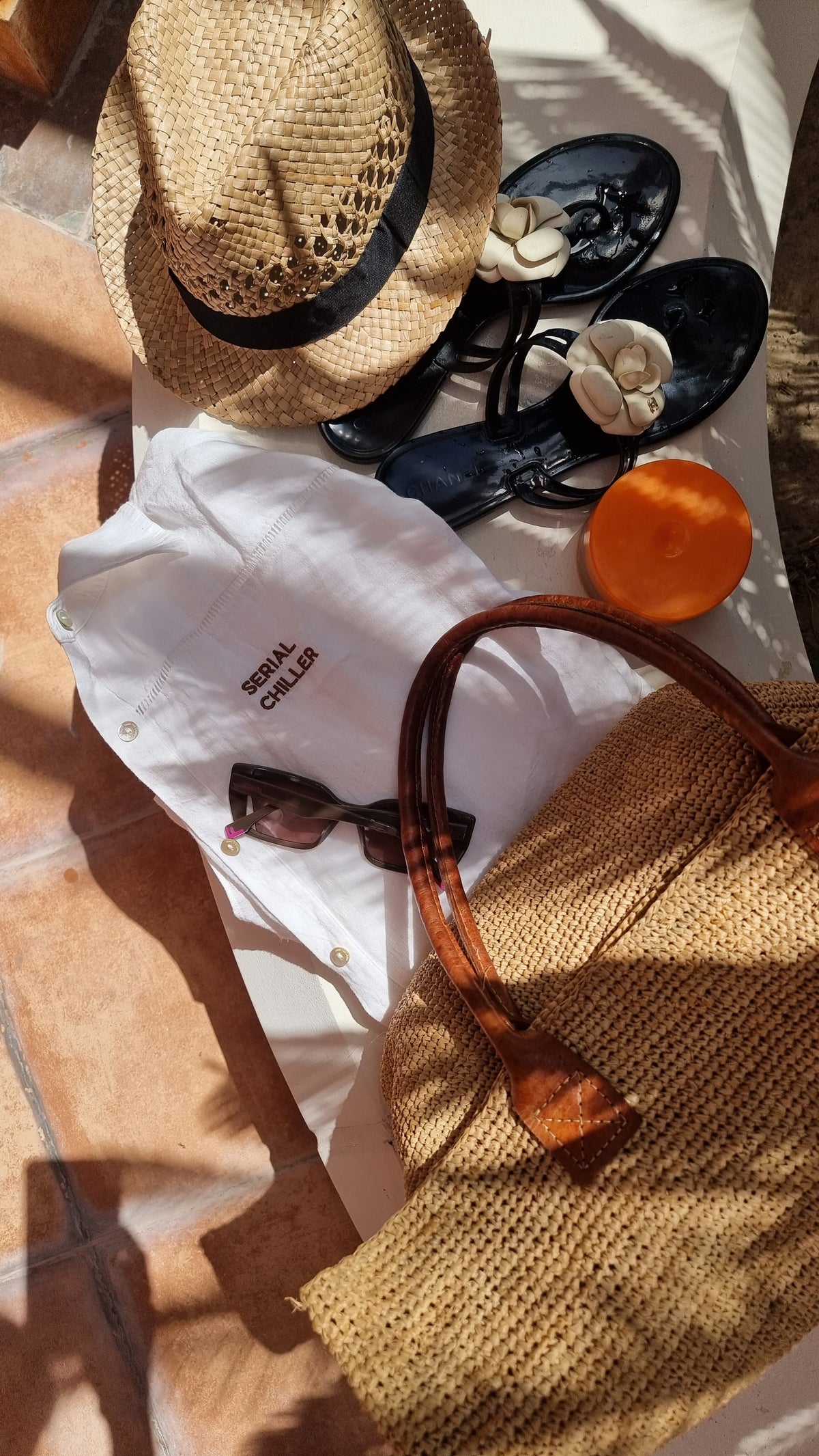 Flat lay of Omni The Label’s white linen shirt with “Serial Chiller” embroidery, styled with a straw hat, sunglasses, black loafers, and a woven beach bag for a summer getaway vibe.UAE, KSA, Qatar