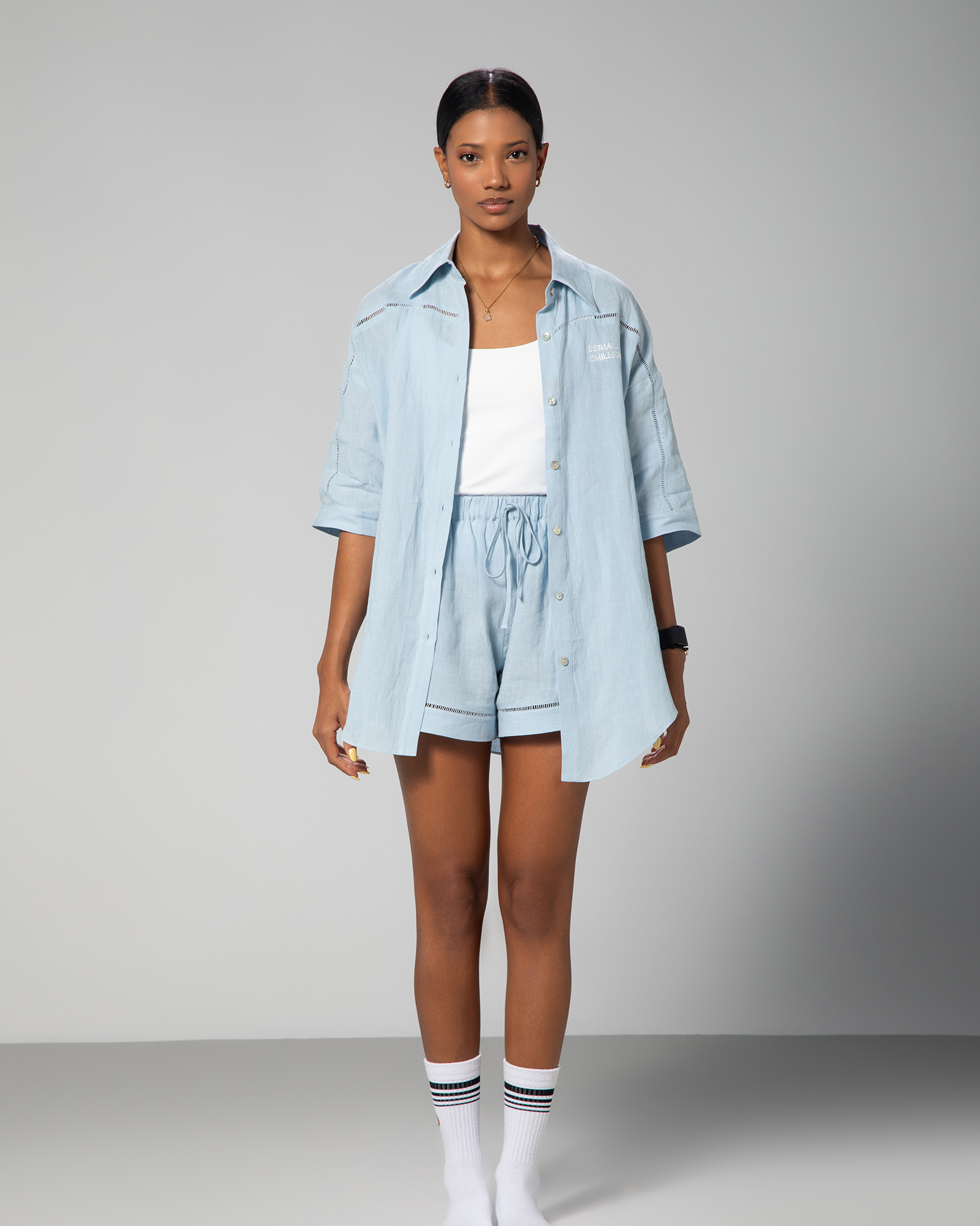 A female model wearing a blue linen shirt   and a matching pair of shorts, standing against a plain background