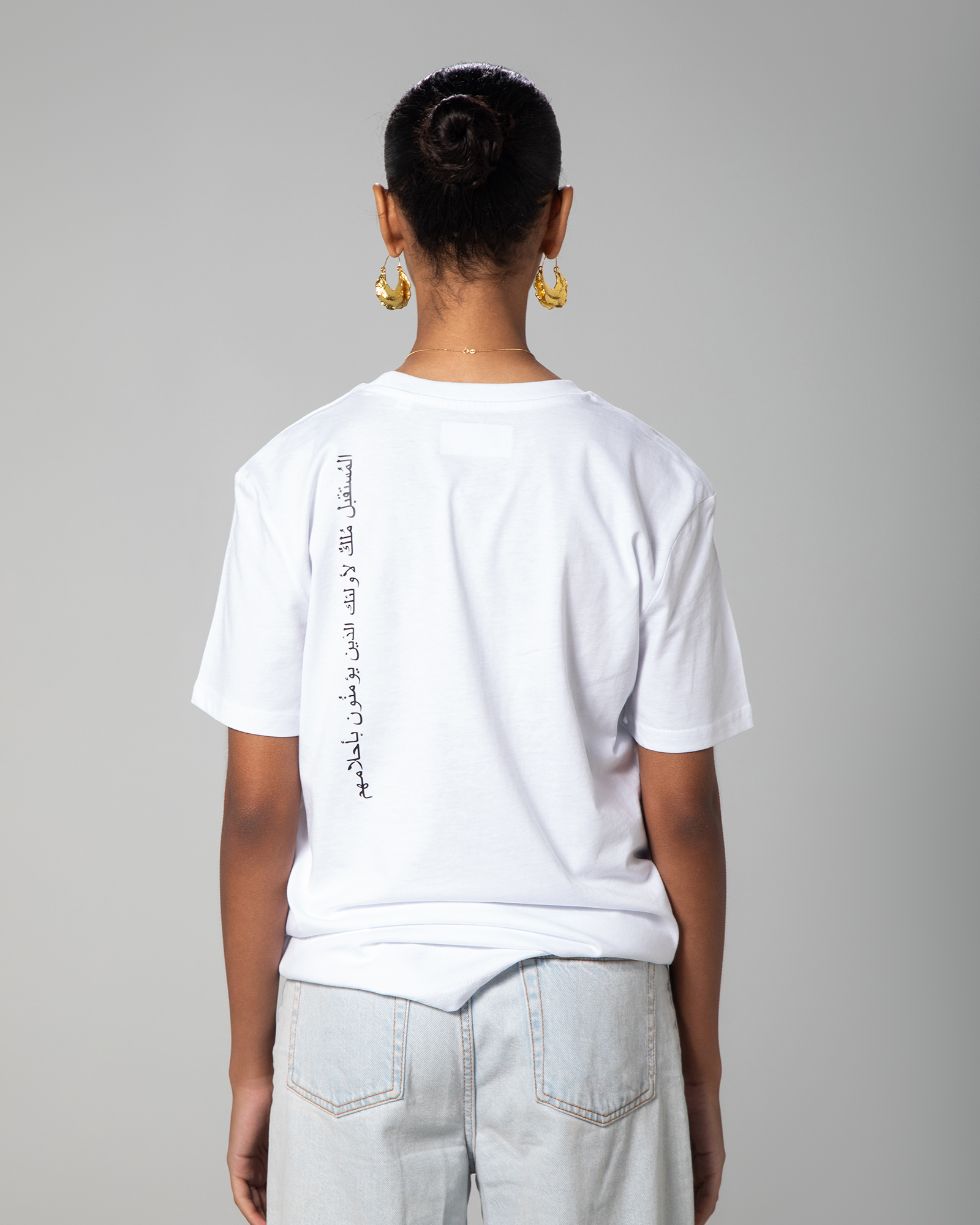 Woman wearing white fitted T-shirt with Arabic calligraphy print on the back, designed by Omni The Label. Modern streetwear  Qatar, UAE, KSA. Sustainable fashion meets culture and comfort.