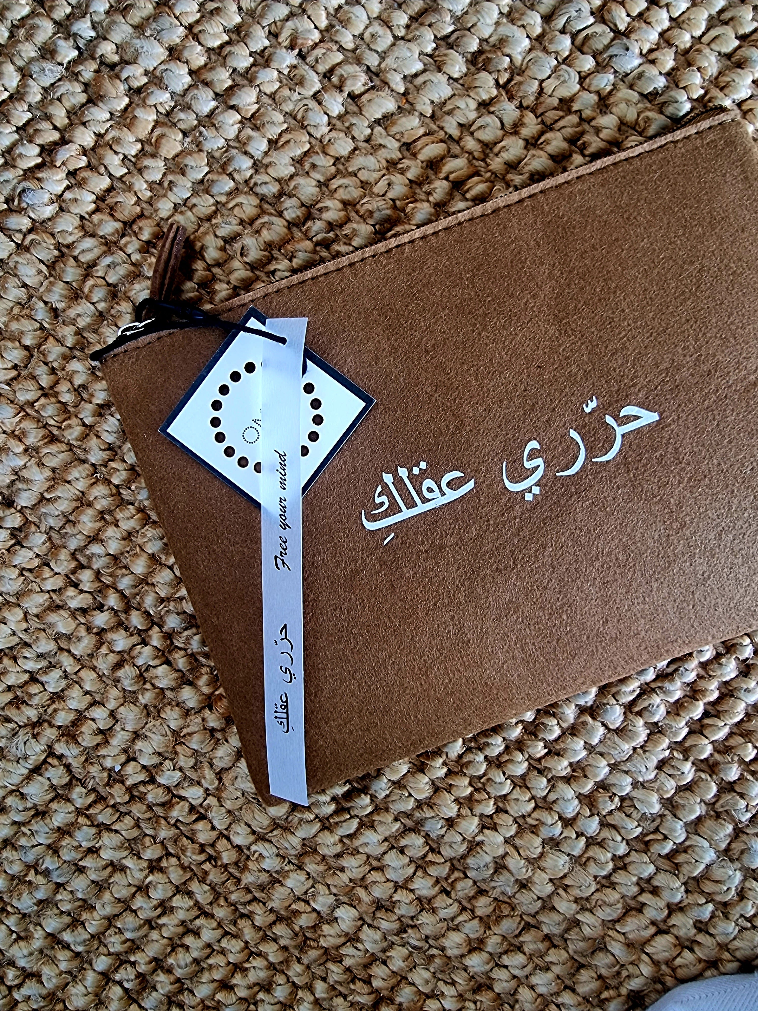 A brown free your mind felt pouch with a printed quote in Arabic lettering, closed with a zipper and a tag attached to it.UAE, KSA, Qatar