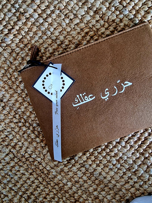 A brown free your mind felt pouch with a printed quote in Arabic lettering, closed with a zipper and a tag attached to it.UAE, KSA, Qatar